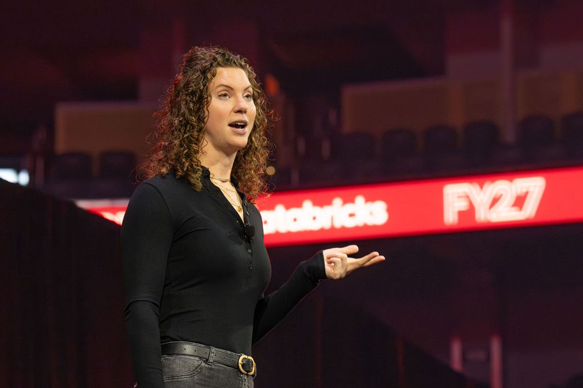 Speaker with curly hair presenting on stage at conference with red branded backdrop