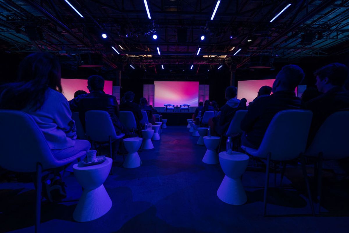 Attendees seated in white chairs facing a stage with pink lighting in a conference venue with blue ambient.