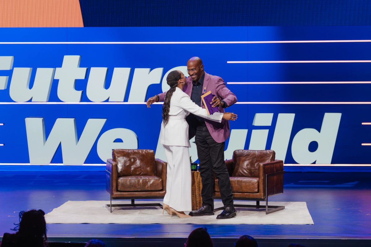 Two speakers embrace on stage with leather chairs and blue backdrop at conference venue