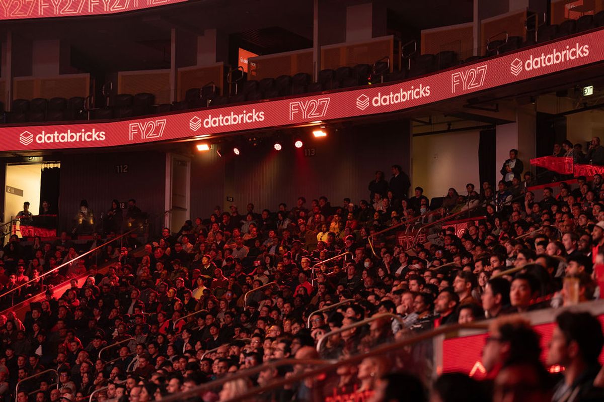 Large crowd of attendees seated in tiered arena seating at Databricks FY27 conference, lit with red lighting