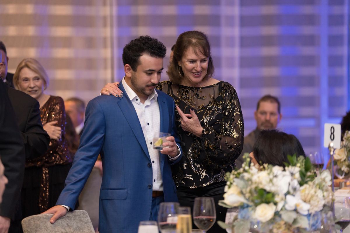 Two people embrace at an elegant dinner event, with formal table settings and floral arrangements visible