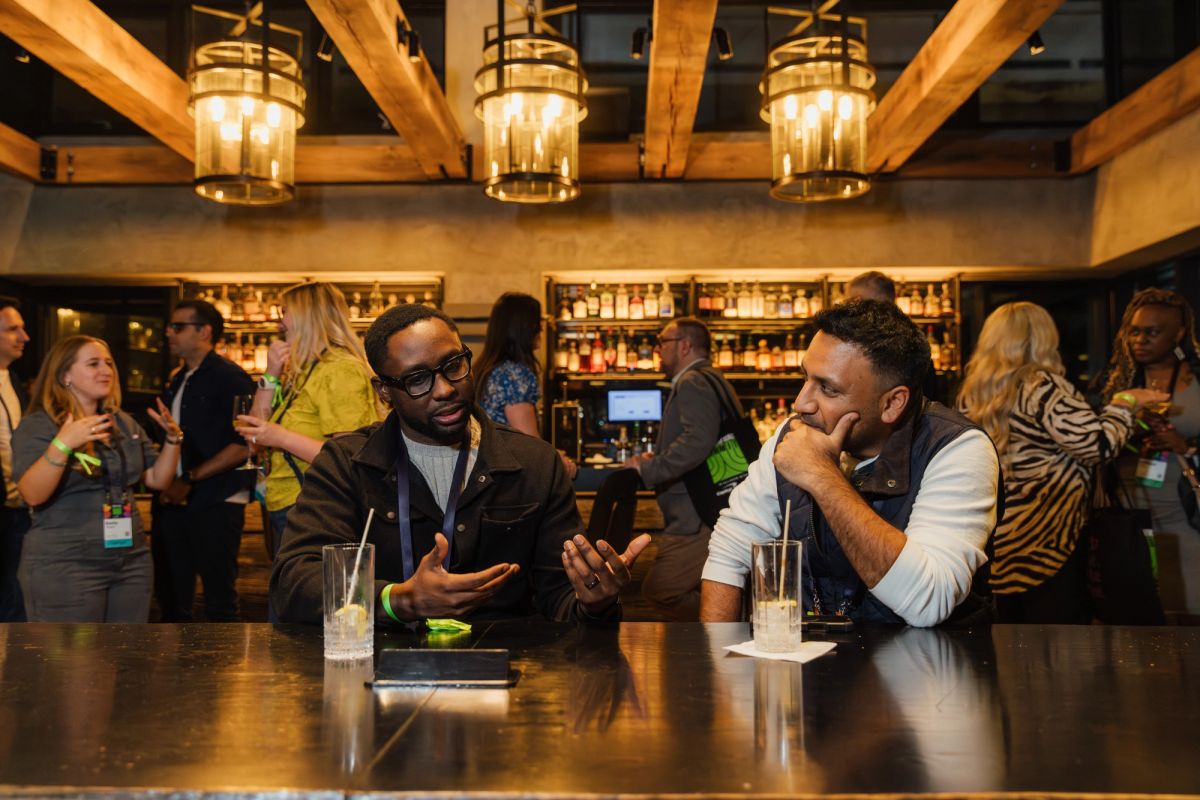 Two men having an animated conversation at a bar with exposed wooden beams and pendant lighting, with other.