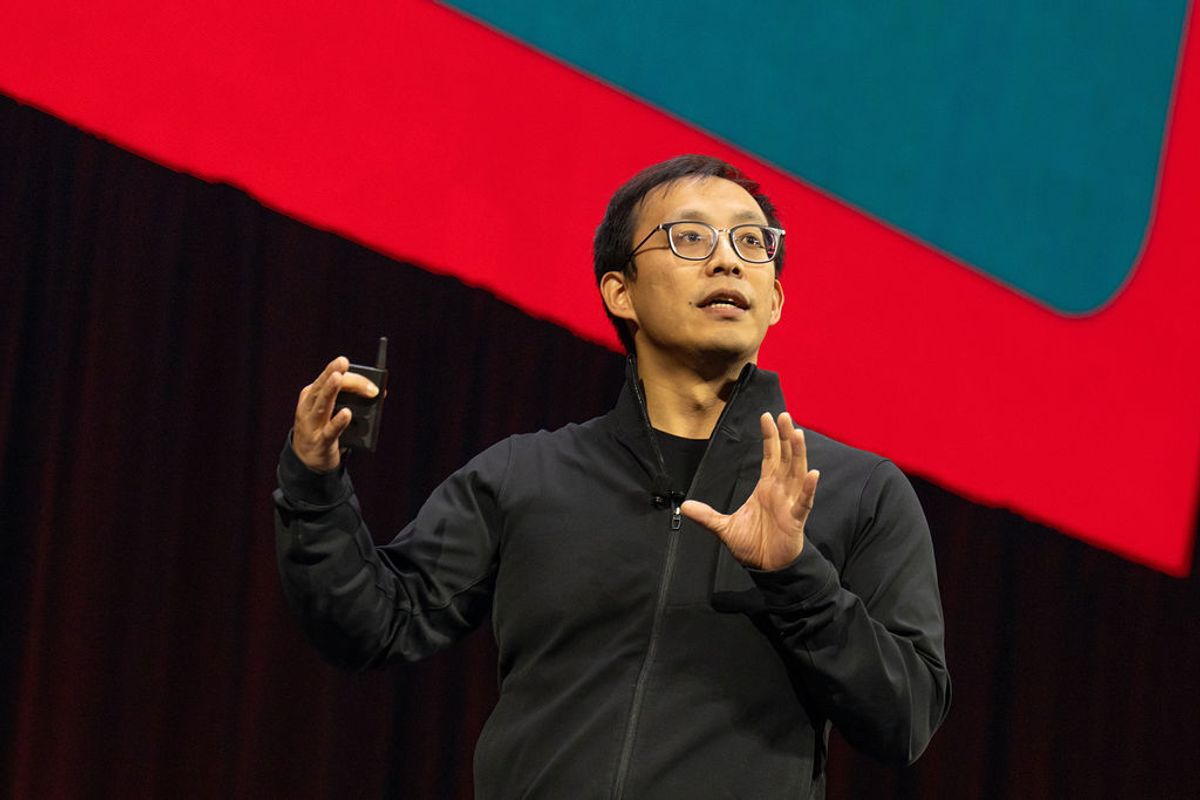 Speaker in black jacket and glasses gesturing while presenting on stage with red lighting in background