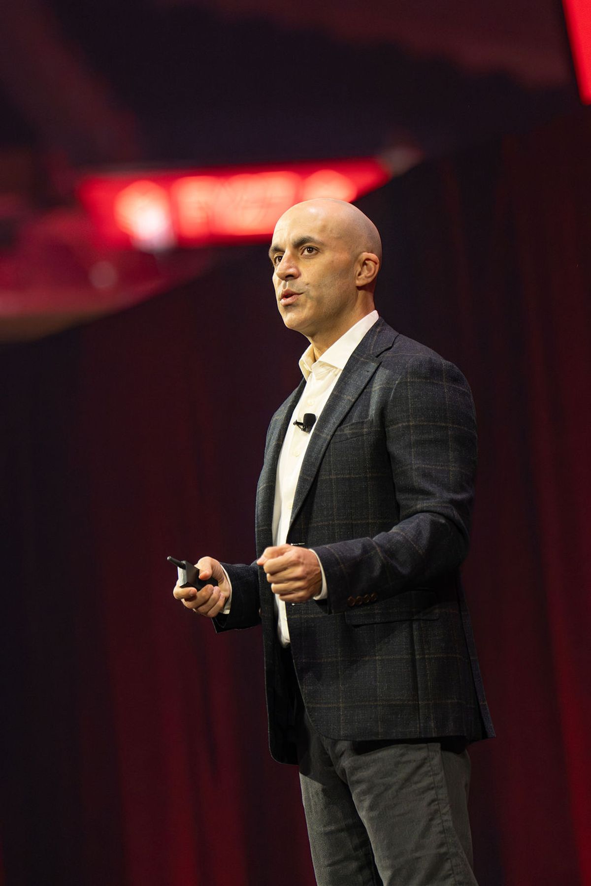 Bald speaker in dark blazer presents on stage with red lighting and blurred backdrop at conference venue
