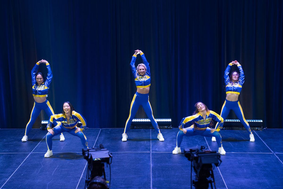 Five cheerleaders in blue and yellow uniforms performing on stage with blue backdrop