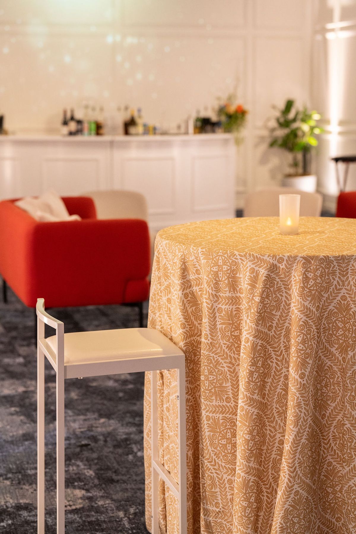 Round table with patterned yellow tablecloth, wooden stool, red armchair, and white kitchen in background