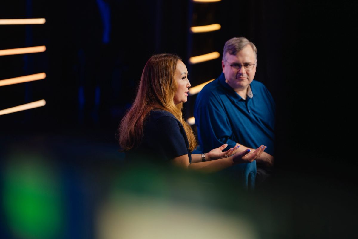 Two speakers on stage in conversation at a conference, with colorful stage lighting in the background