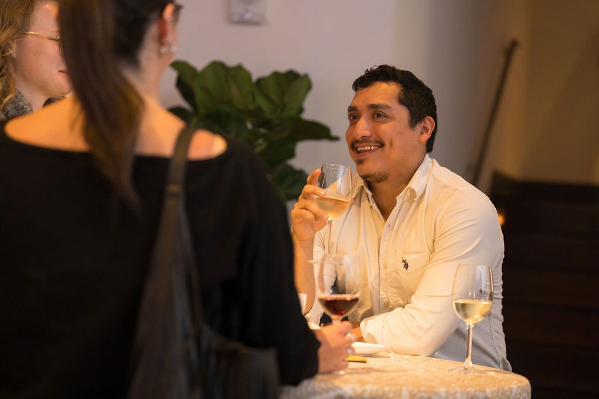 Man in white shirt holds wine glass while networking with attendees at indoor reception event.