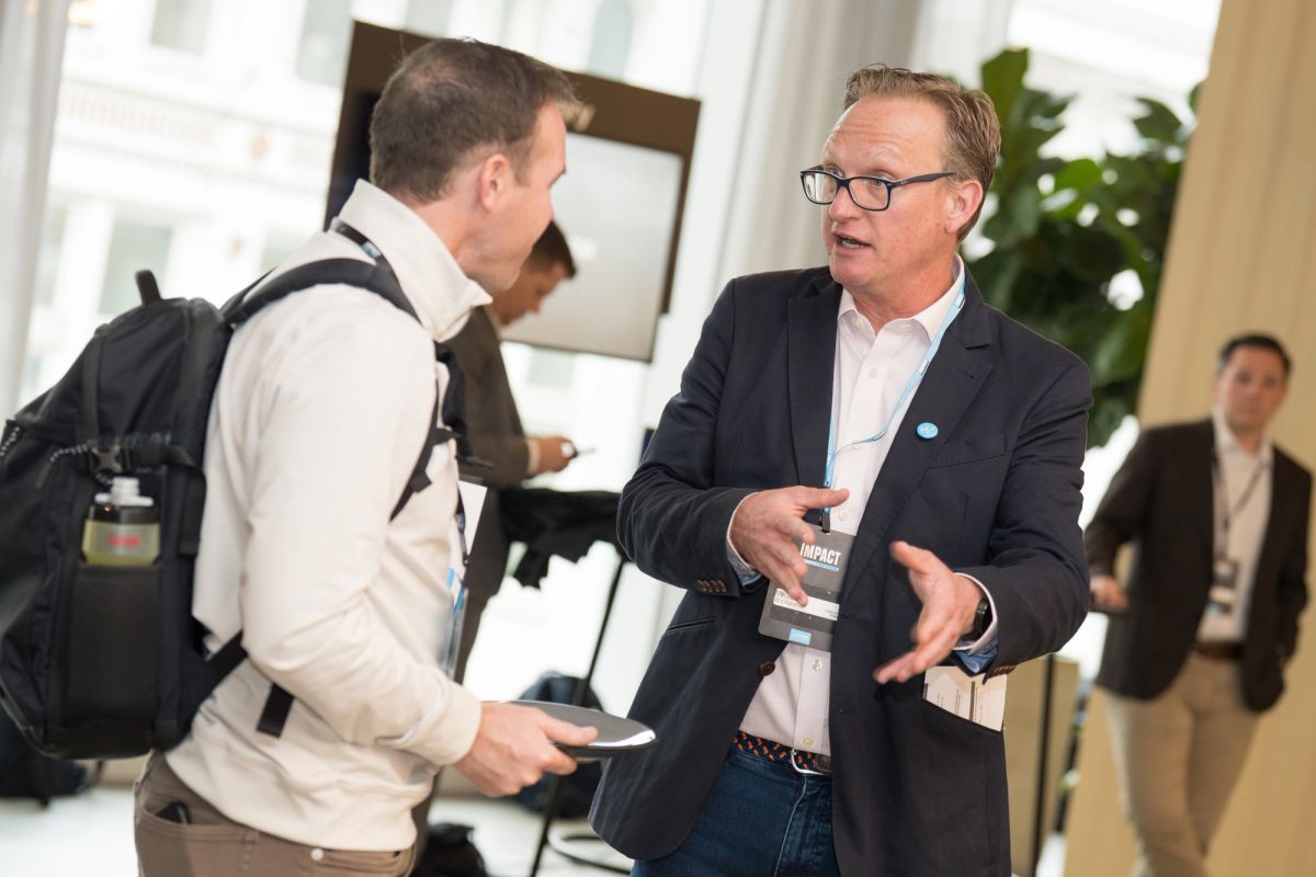 Two men in business attire having a conversation at a conference, one wearing glasses and holding materials.