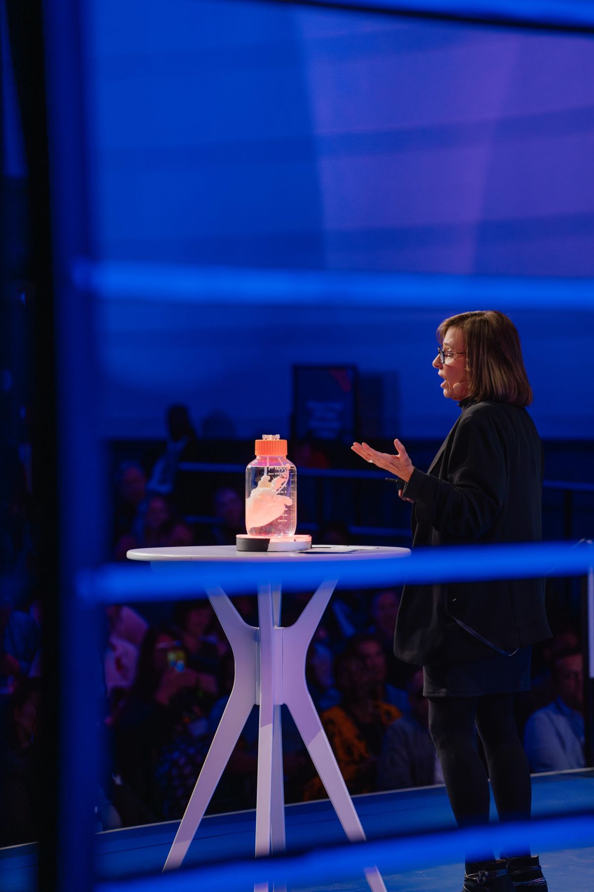 Speaker in black presenting beside white table with water bottle at conference venue with blue lighting