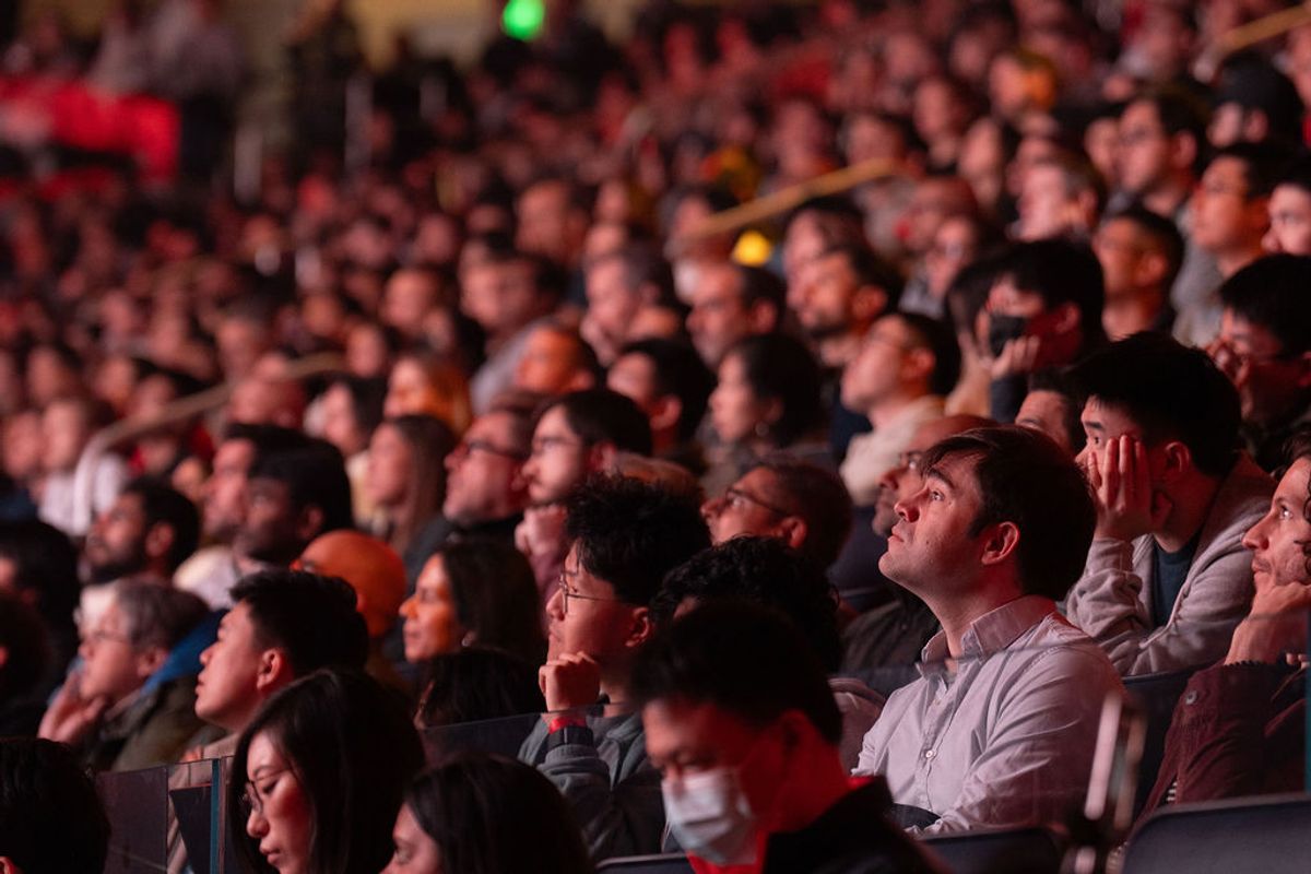 Two men sitting in audience seats looking up, with blurred crowd visible in background under red lighting