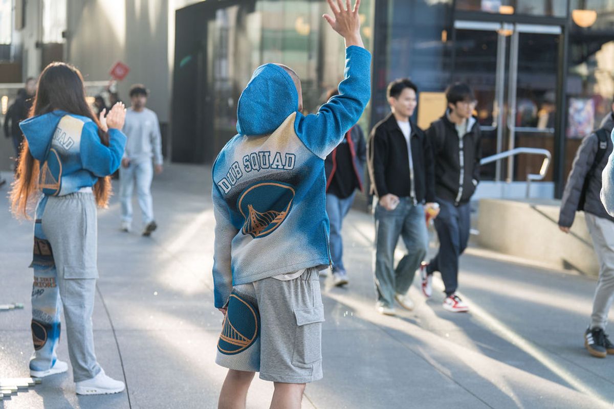 Person in blue hoodie waves while walking through outdoor plaza area with other people nearby