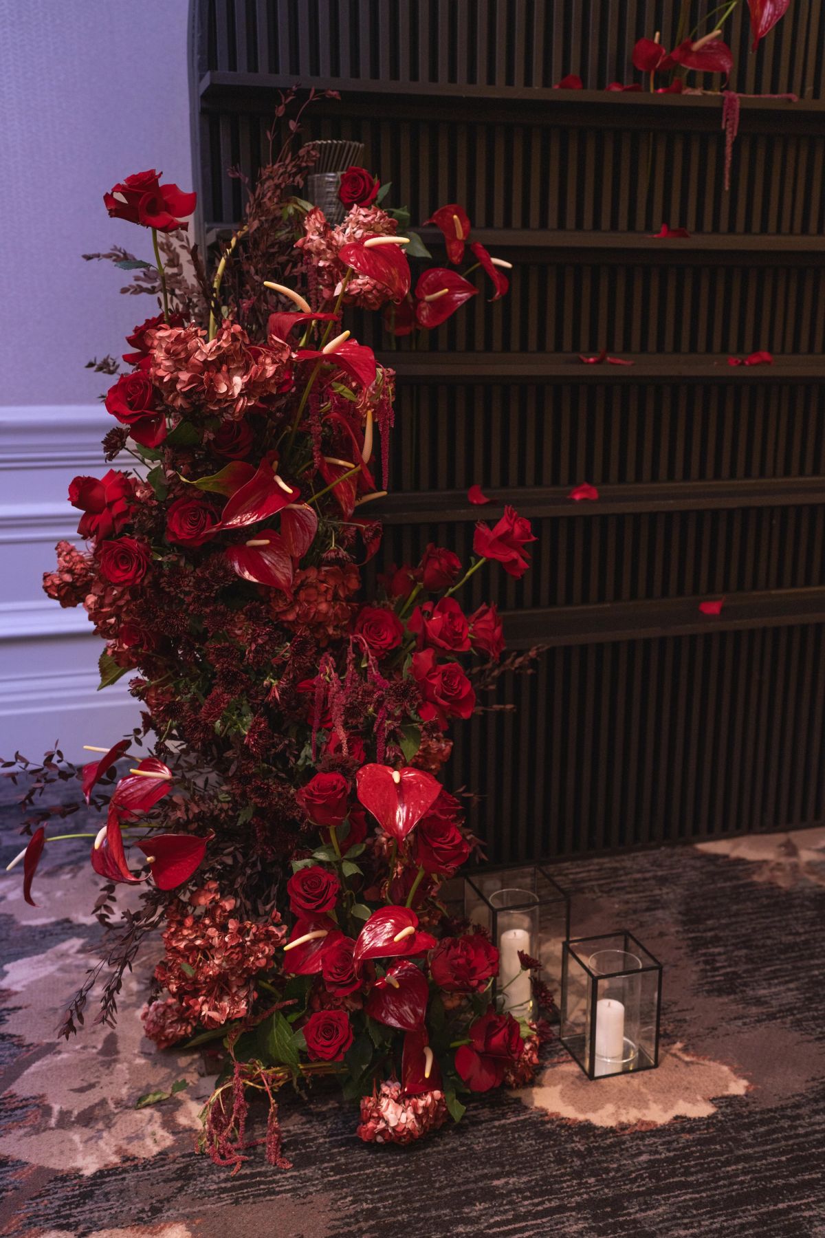 Large red floral arrangement with roses and anthuriums beside wooden stairs, with glass candle holders nearby