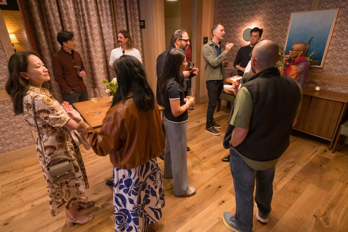 Group of people socializing in a warmly lit room with wooden floors, curtains, and wall art during an event