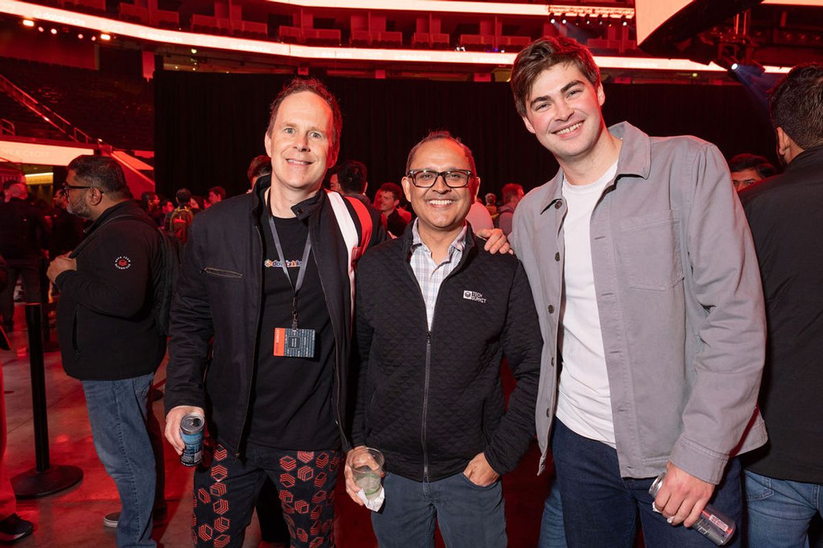 Three men pose together smiling at a conference event with red lighting and attendees in background