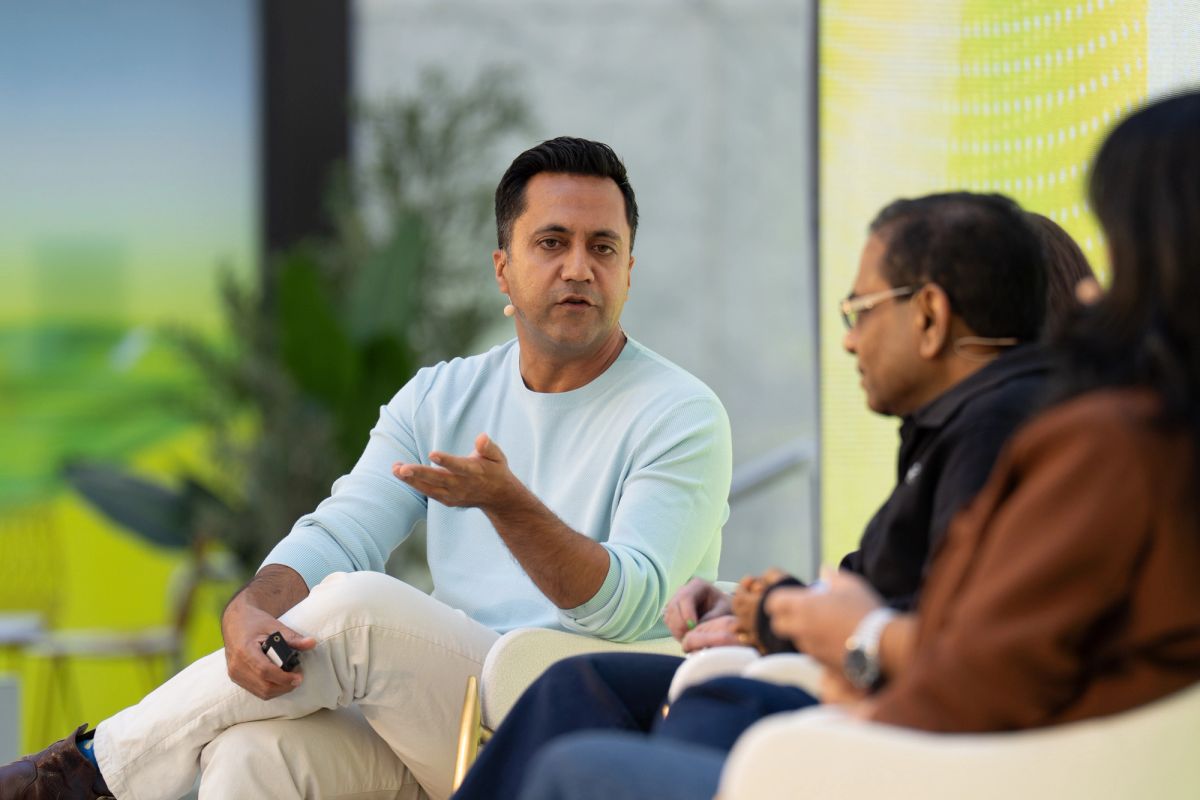 Panelist in light blue sweater gestures while speaking to two seated attendees at a conference.