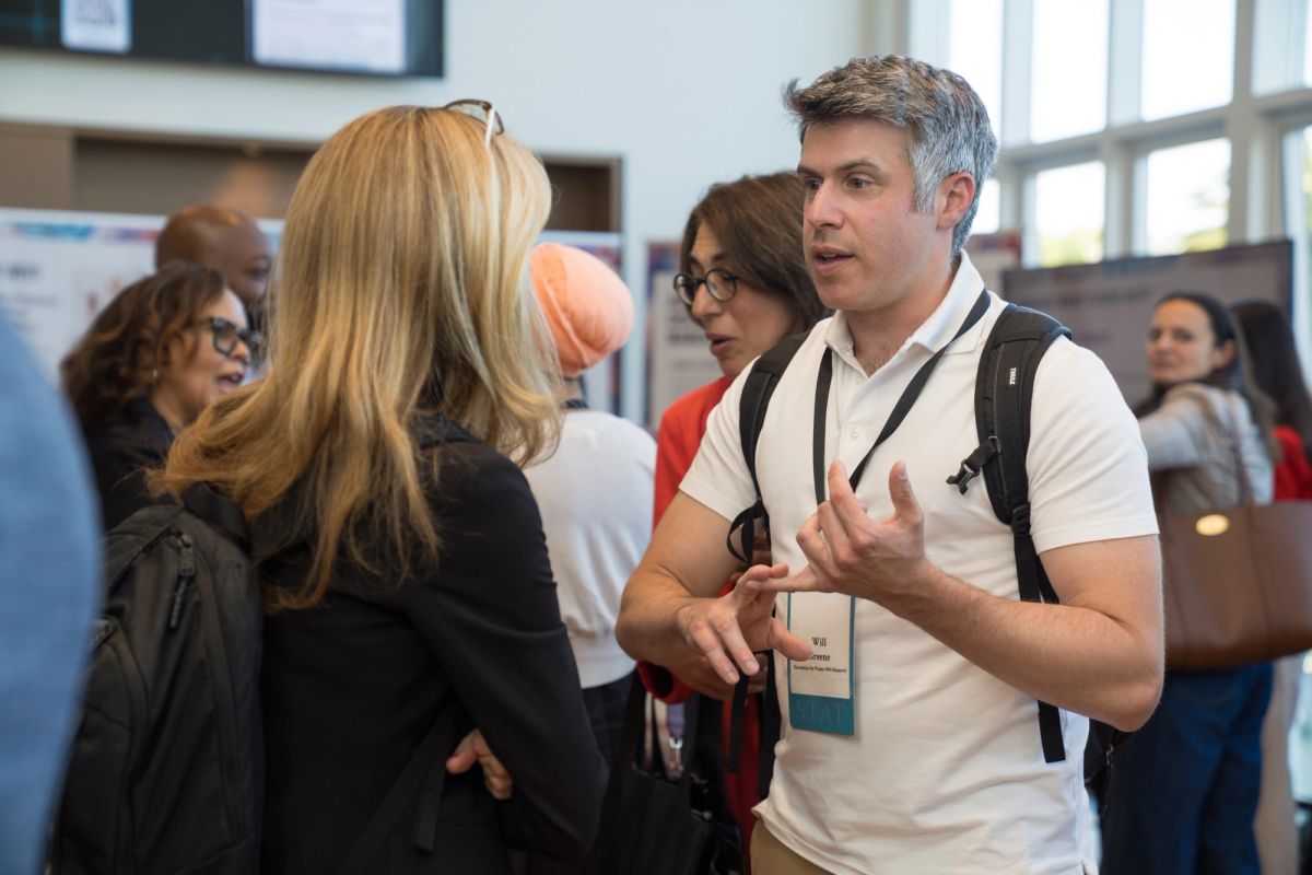Conference attendees networking and conversing in a venue with windows and displays in the background.