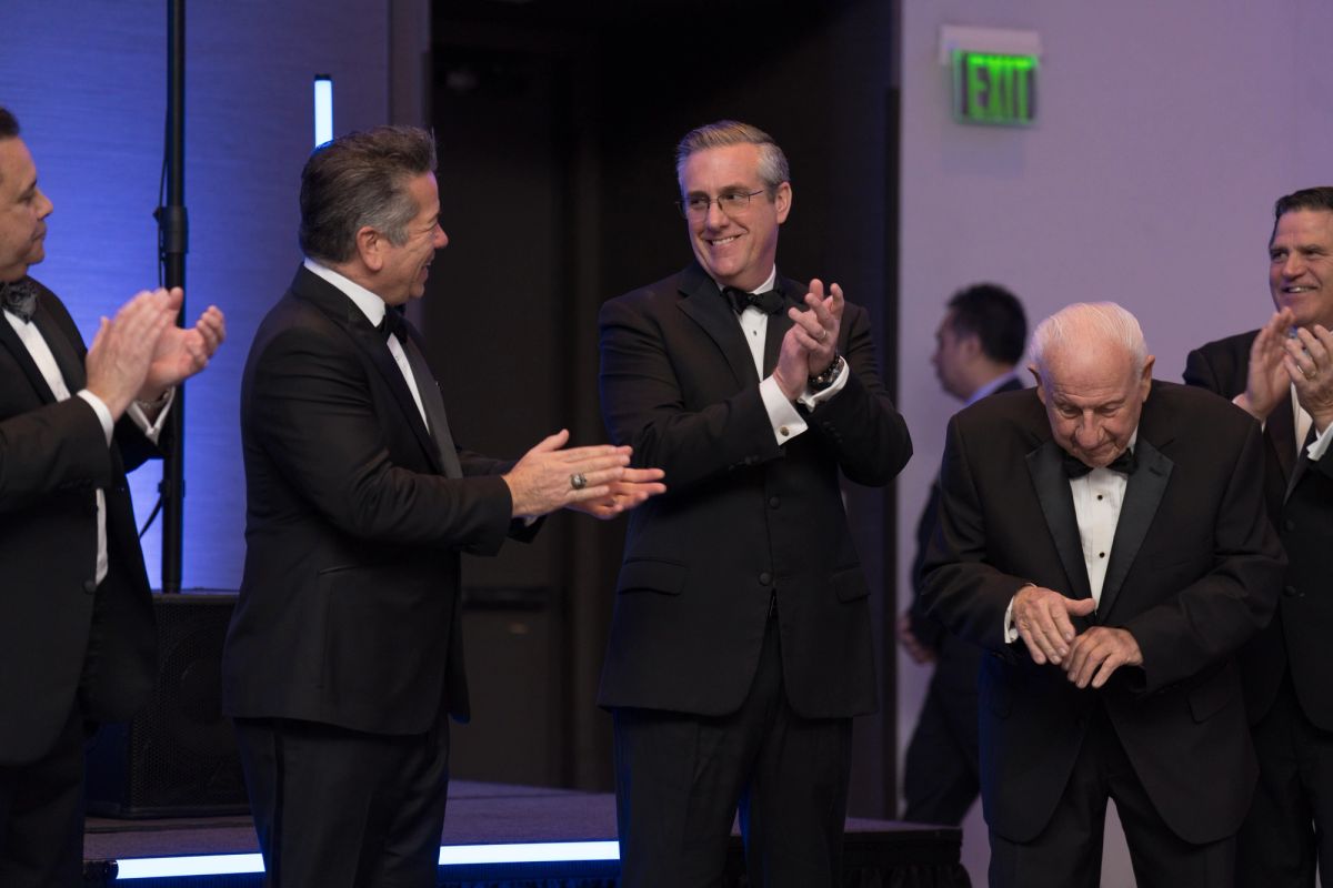 Men in tuxedos applauding as an older gentleman in formal wear takes a bow at a gala event.