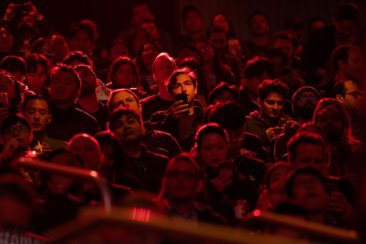 Audience members seated in red lighting at an indoor event, some holding phones to record or take photos.