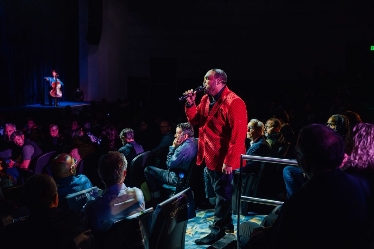 Speaker in red shirt with microphone performing for seated audience in dark theater venue
