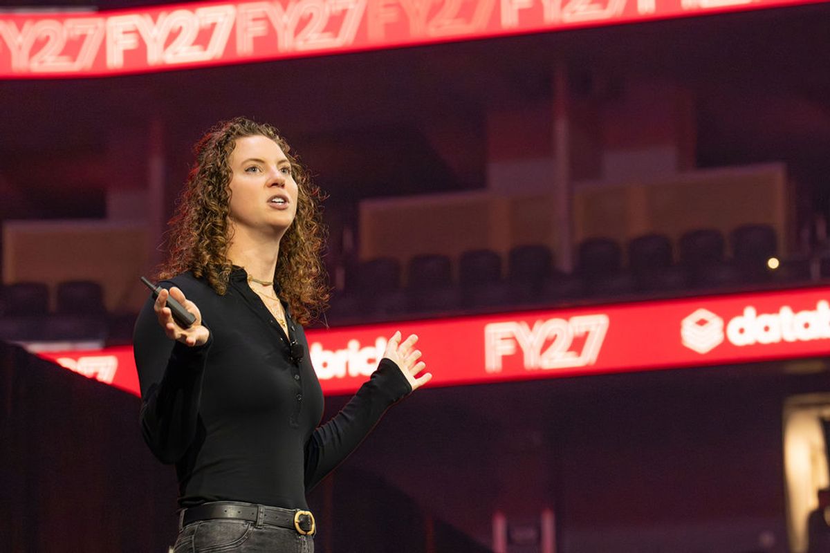 Speaker with curly hair presenting on stage at conference venue with red digital signage in background