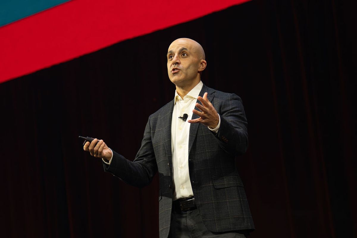 Bald speaker in gray suit presenting on stage with microphone, gesturing with one hand while holding device.