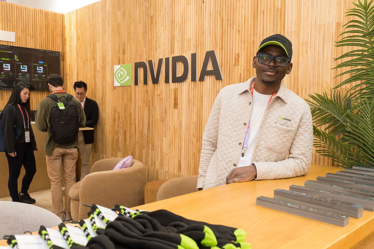 Attendee in cream jacket and cap stands at NVIDIA booth table with merchandise while other conference.