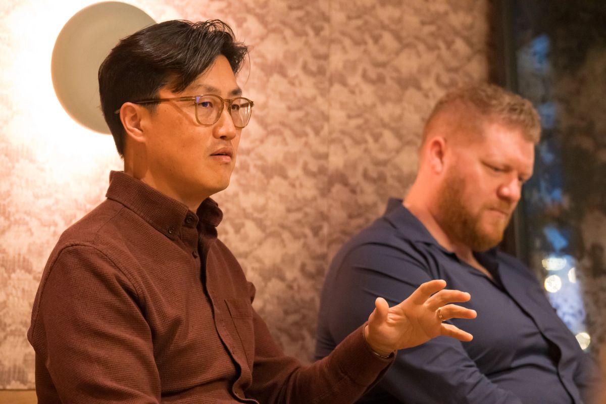 Two panelists during discussion, one gesturing while speaking, seated against textured wall backdrop