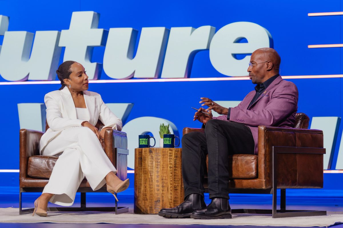 Two speakers in conversation on stage, woman in white suit and man in purple jacket, seated in leather chairs.
