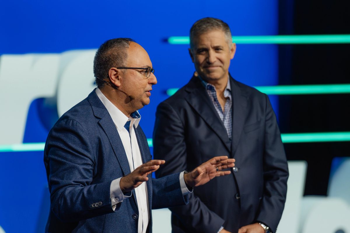 Two business executives on stage, one gesturing while speaking, the other listening, with blue and green.