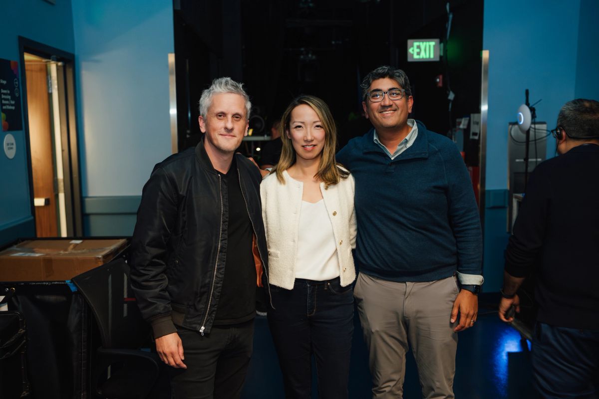 Three people pose together in a theater venue with blue lighting and an exit sign visible in the background.