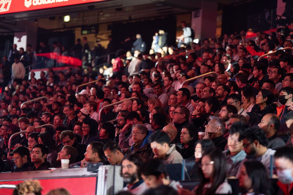 Large crowd of attendees seated in arena-style seating at an indoor conference event, bathed in red lighting