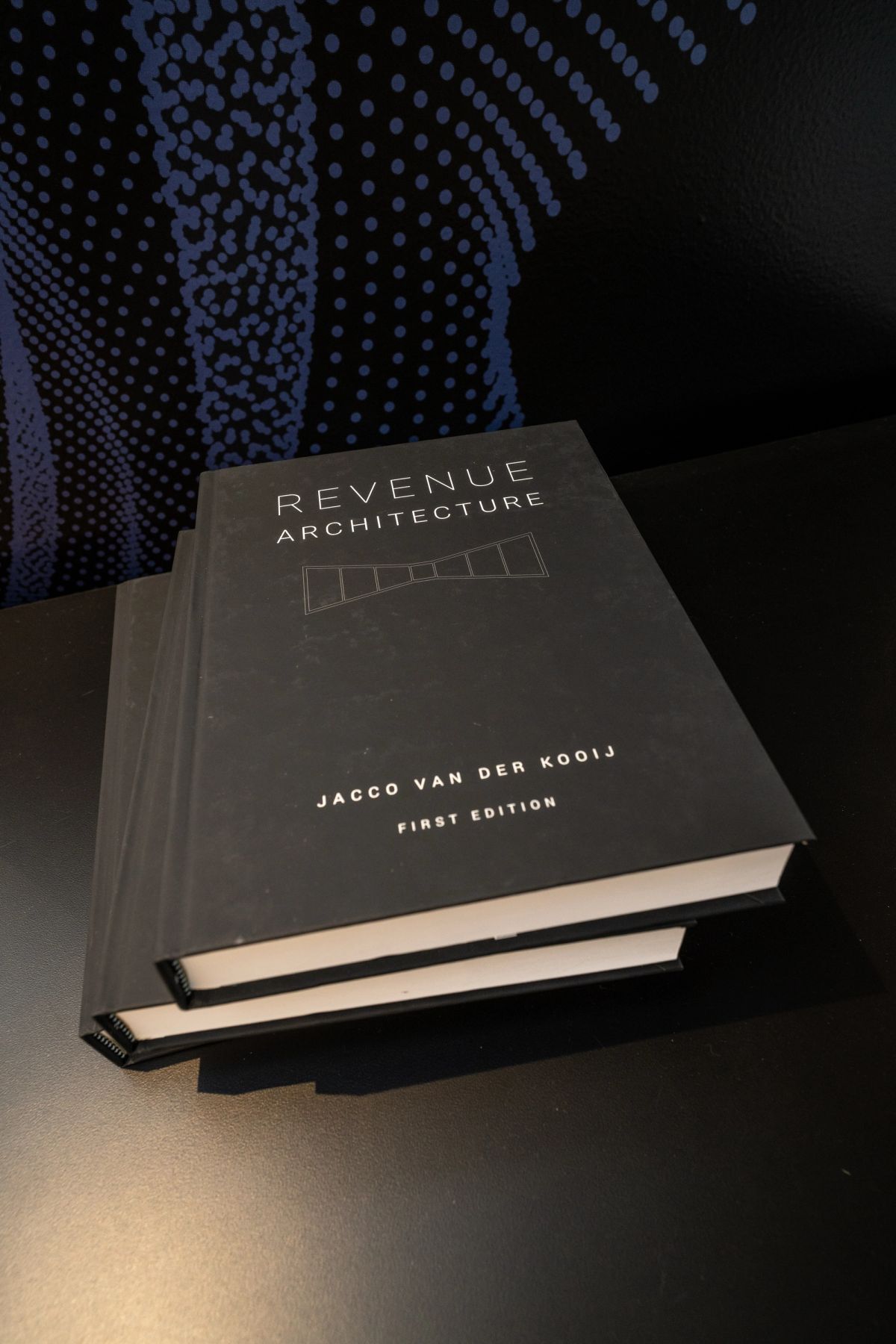 Stack of books titled Revenue Architecture by Jacco van der Kooij on a table with blue dotted backdrop