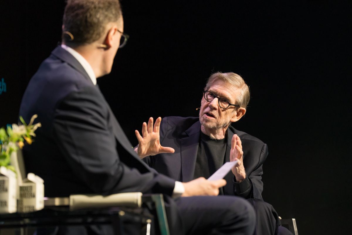 Two men in suits having a conversation on stage, one gesturing with his hands while speaking to the other.
