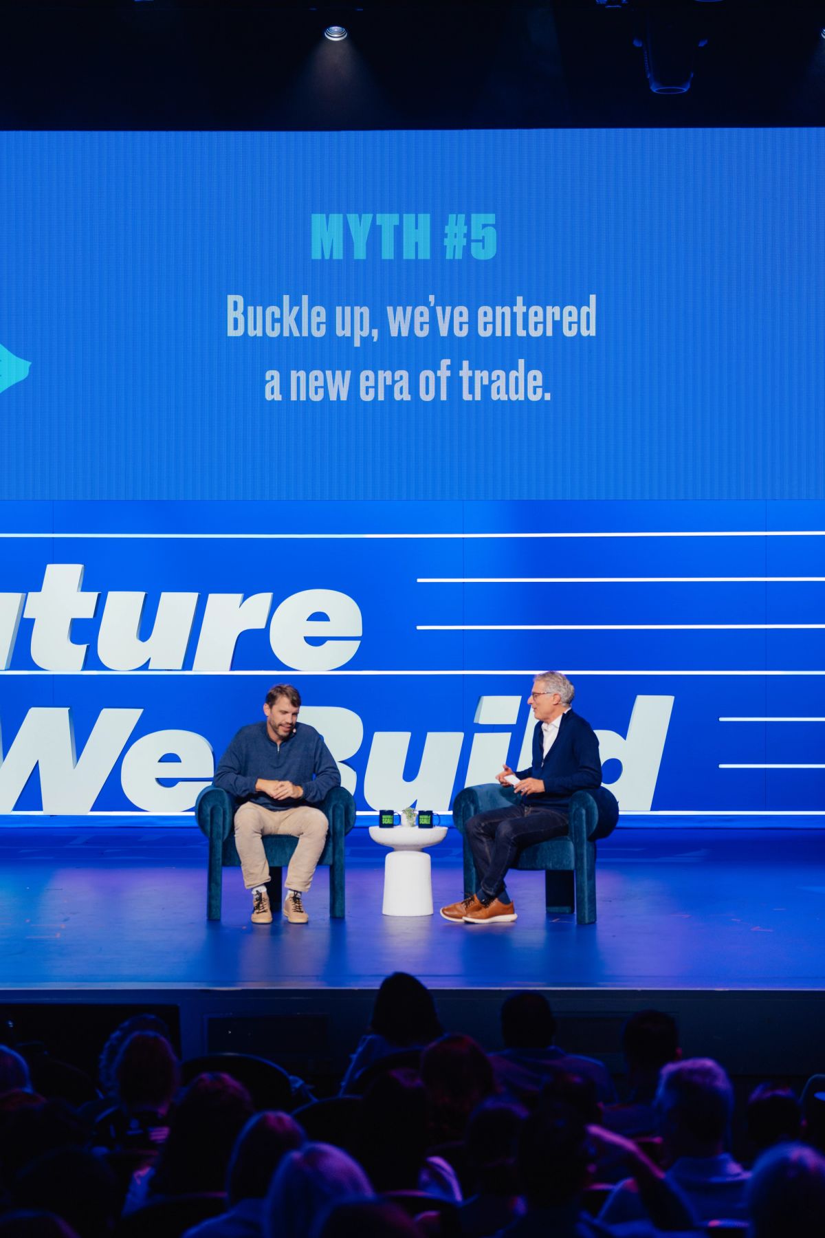 Two speakers in conversation on stage with blue backdrop displaying "MYTH #5 Buckle up, we've entered a new.