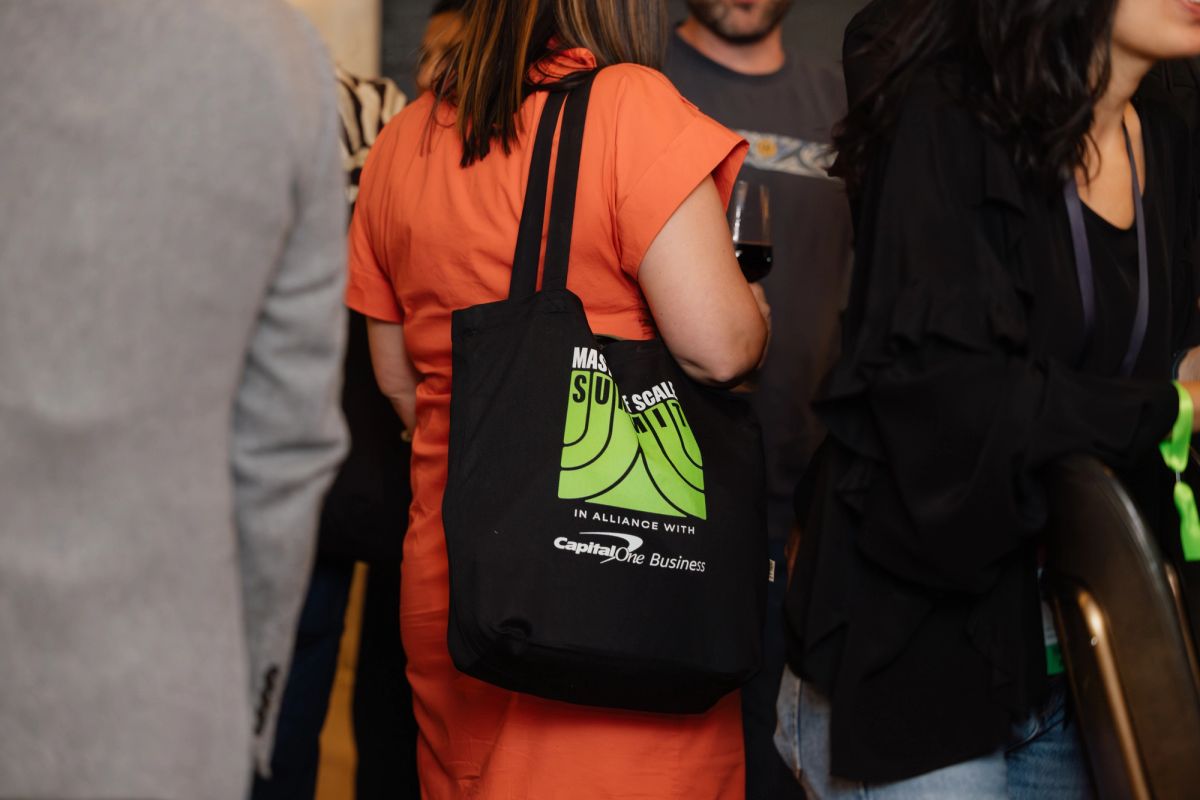 Attendees at conference, person in orange shirt carrying black tote bag with green logo design