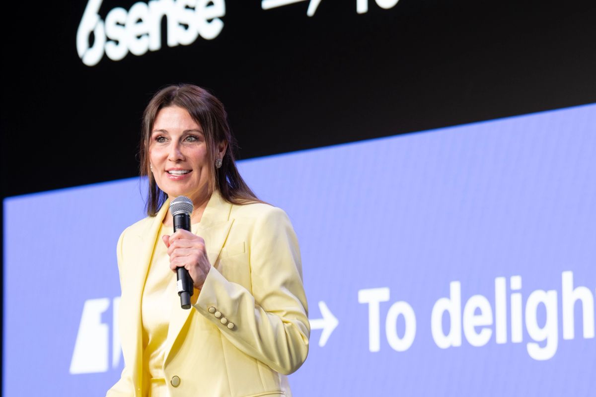 Female speaker in yellow blazer holds microphone on stage with purple backdrop displaying partial text