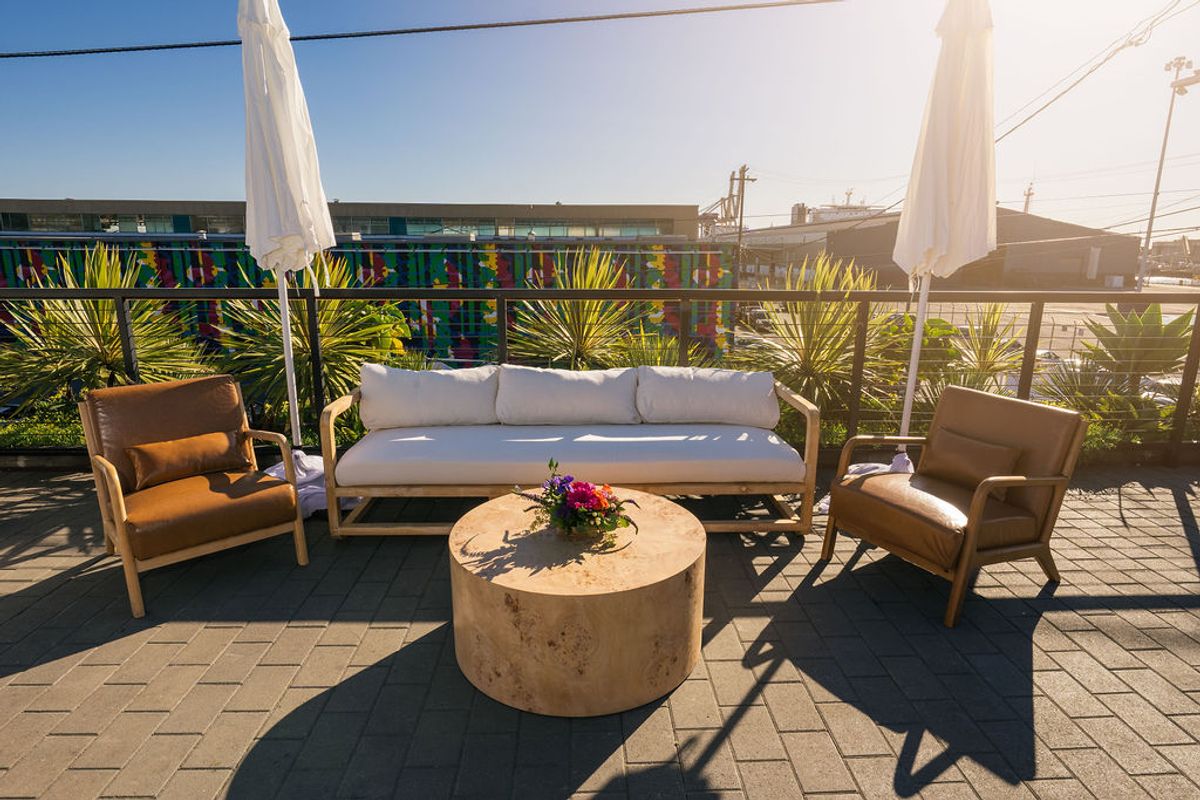 Outdoor rooftop terrace with white sofa, two beige chairs, round coffee table, and palm plants under umbrellas