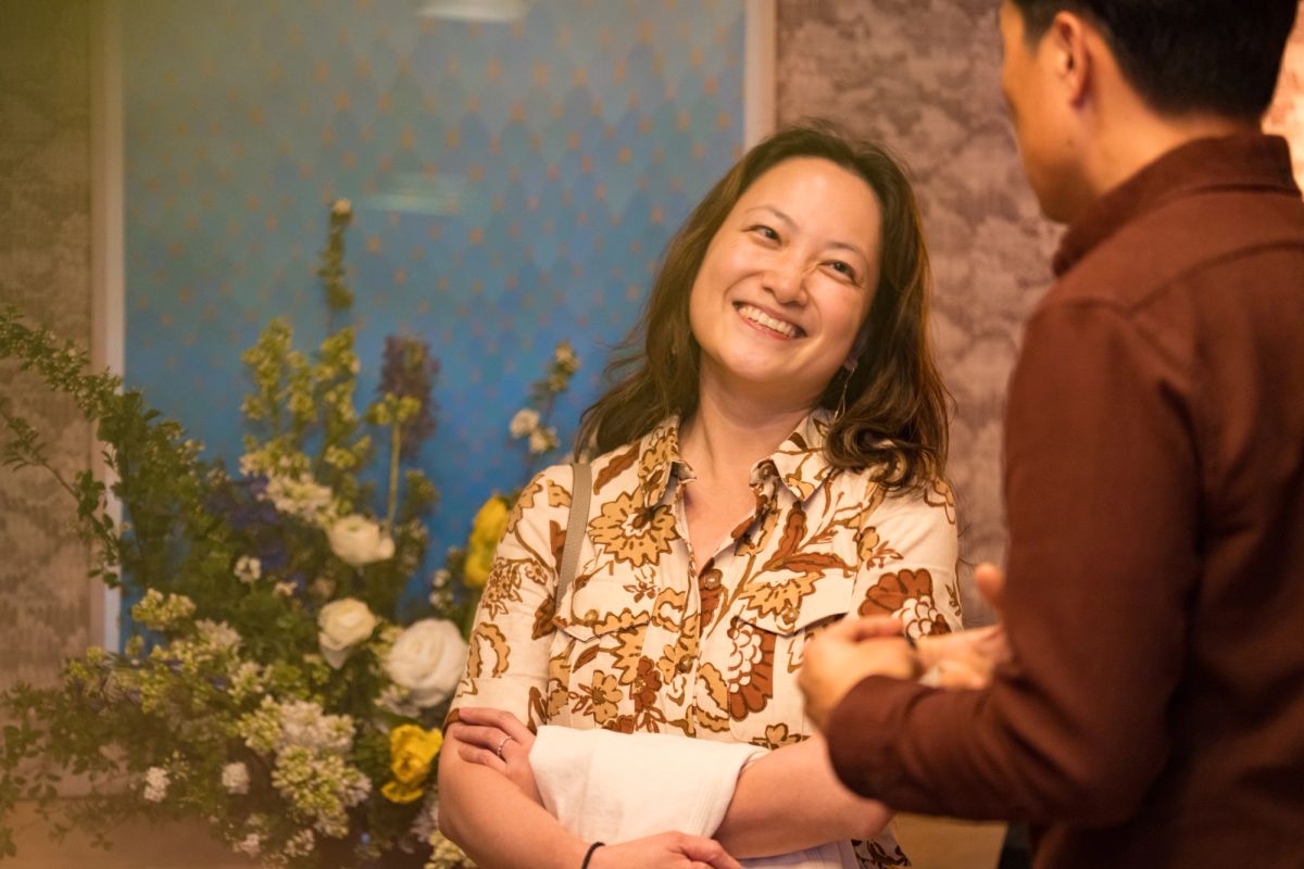 Woman in floral shirt smiling while speaking with man in brown shirt at indoor event with flowers nearby
