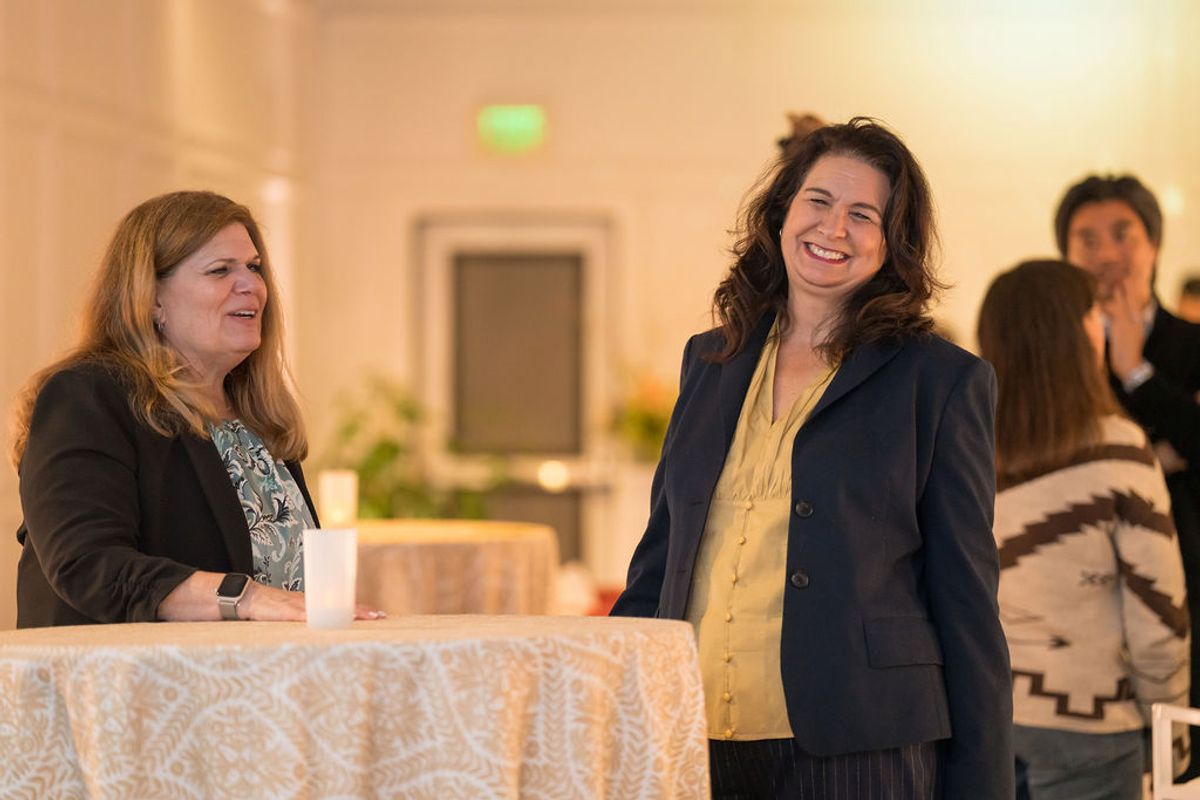 Two businesswomen networking at reception, one standing behind table with white lace tablecloth, both smiling