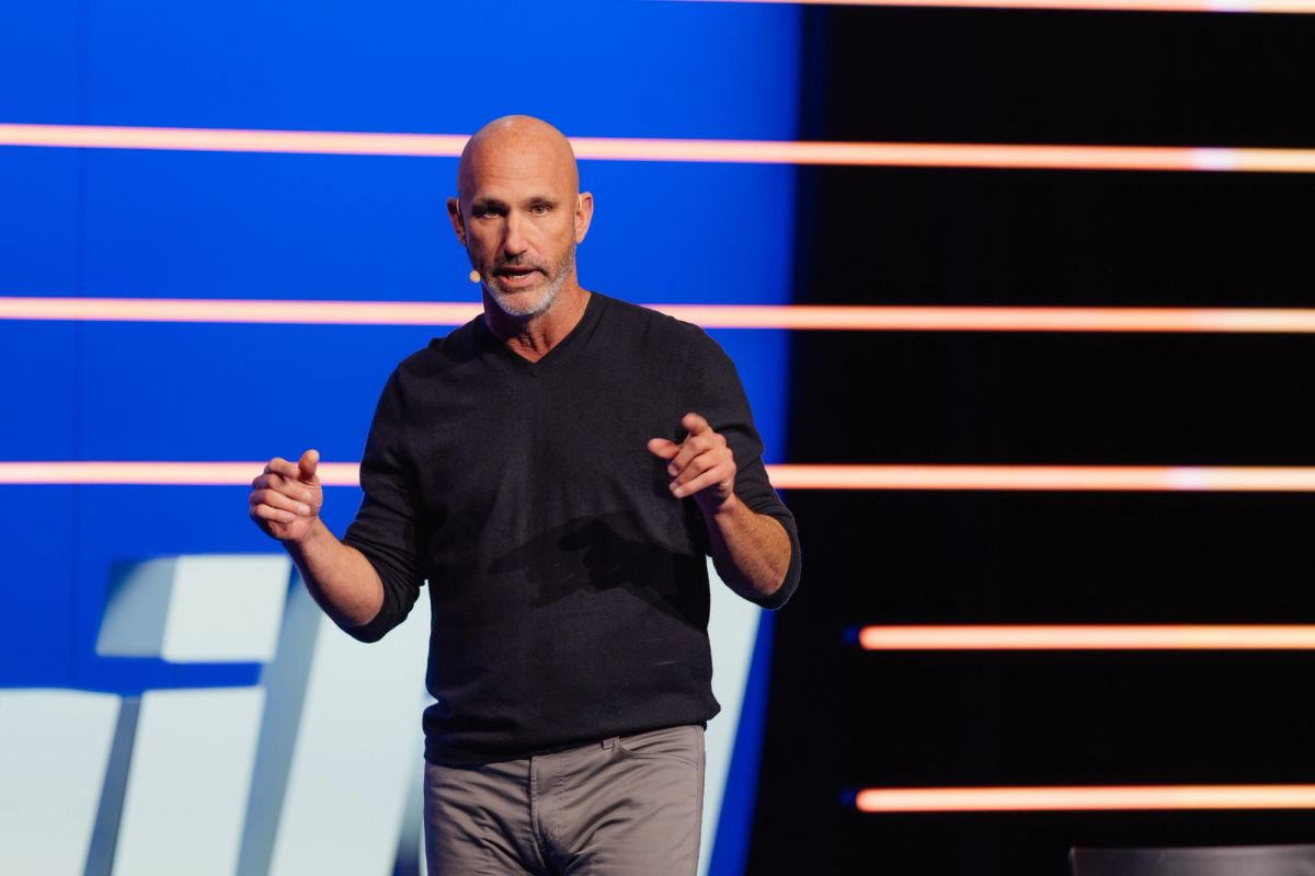 Bald speaker in black sweater gesturing with both hands on stage with blue and black striped backdrop