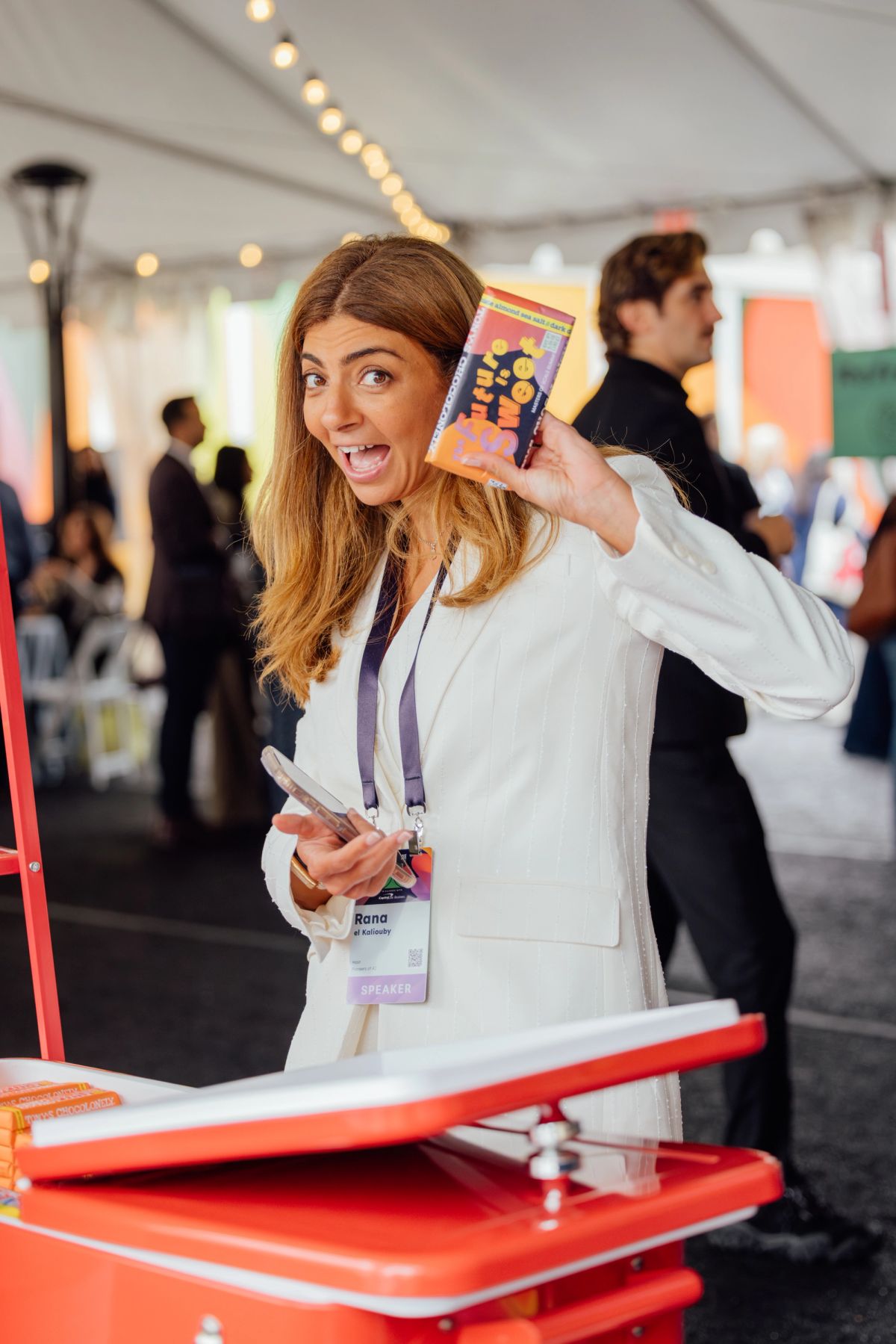Woman in white blazer holds up book while smiling excitedly at conference event under tent with string lights