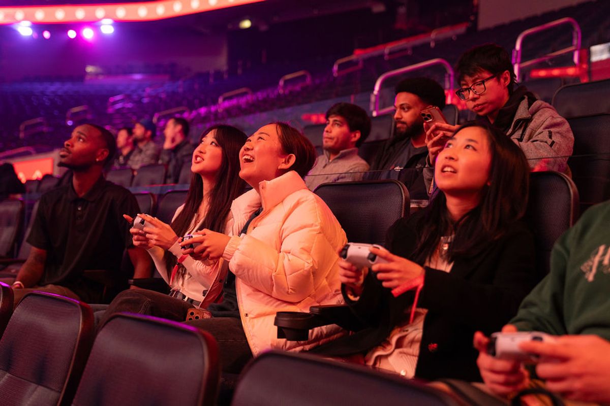 Audience members with gaming controllers sitting in theater-style seating under colorful stage lighting