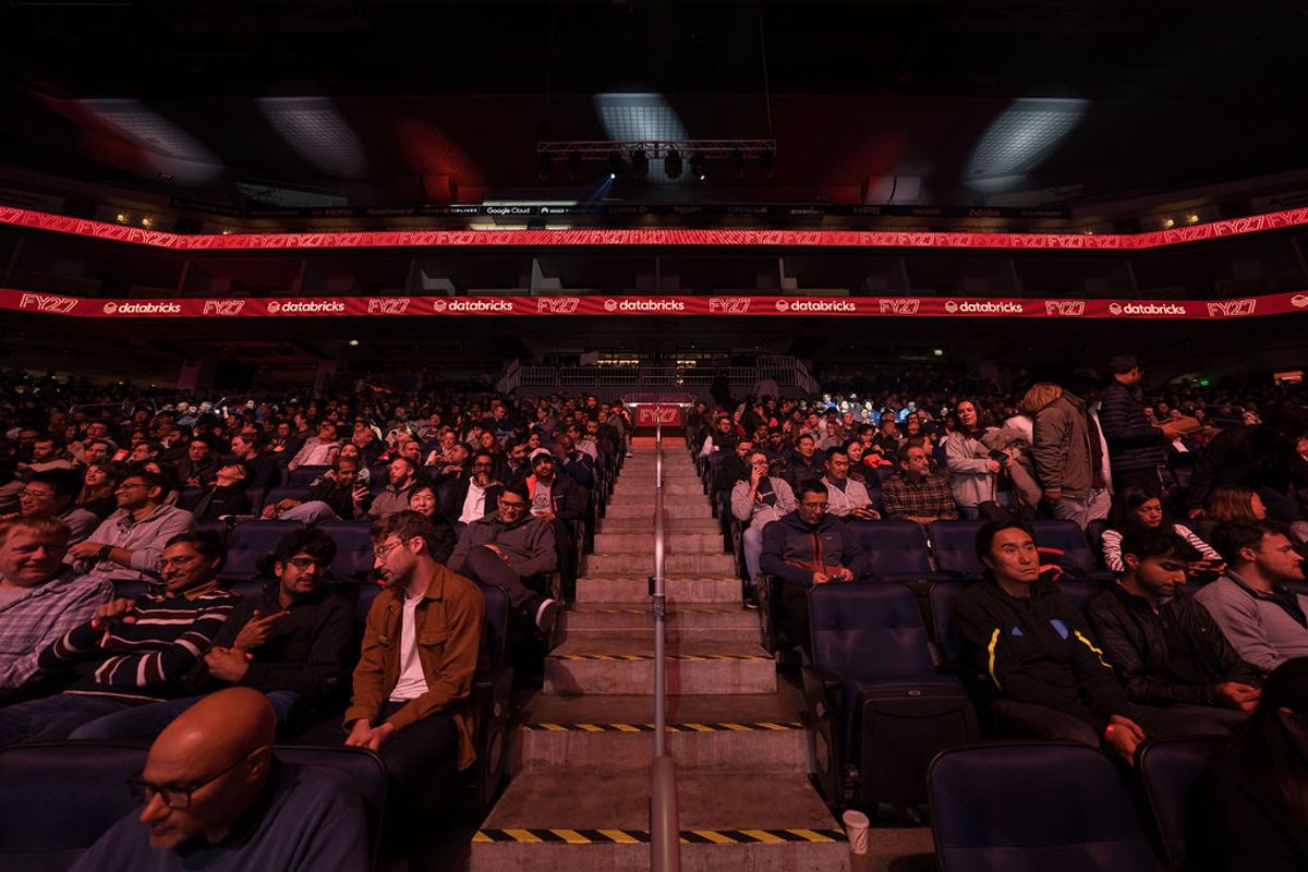 Audience members seated in tiered rows of blue chairs in a large arena venue with red lighting and screens