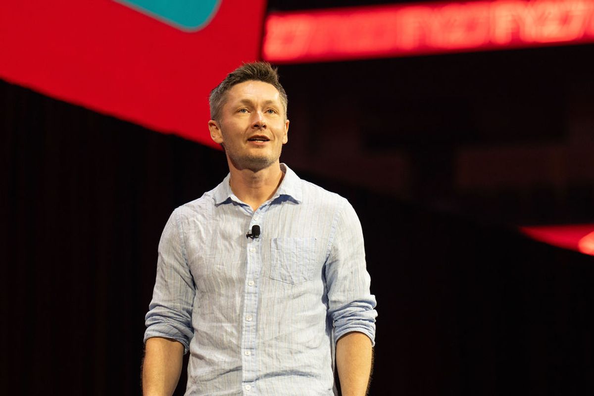 Speaker in light blue shirt presenting on stage with red and black geometric backdrop