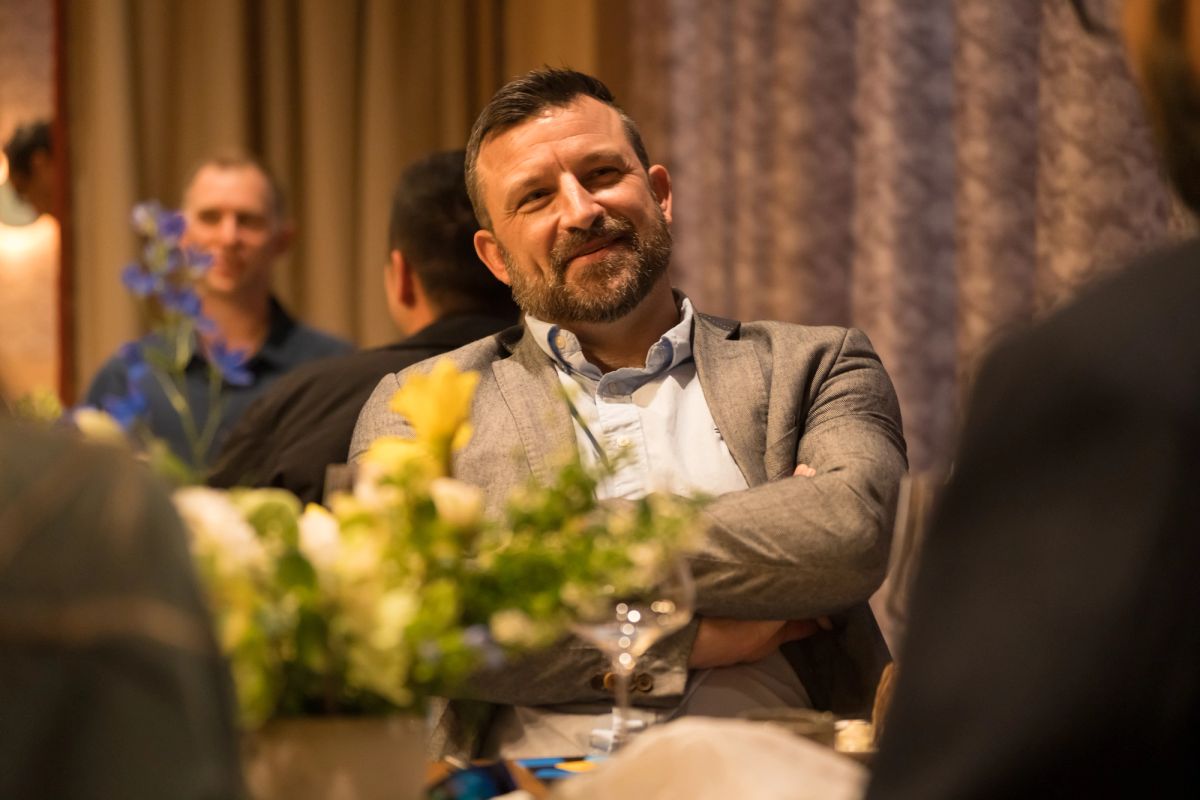 Smiling bearded man in gray blazer sits at panel discussion table with flowers and water glass