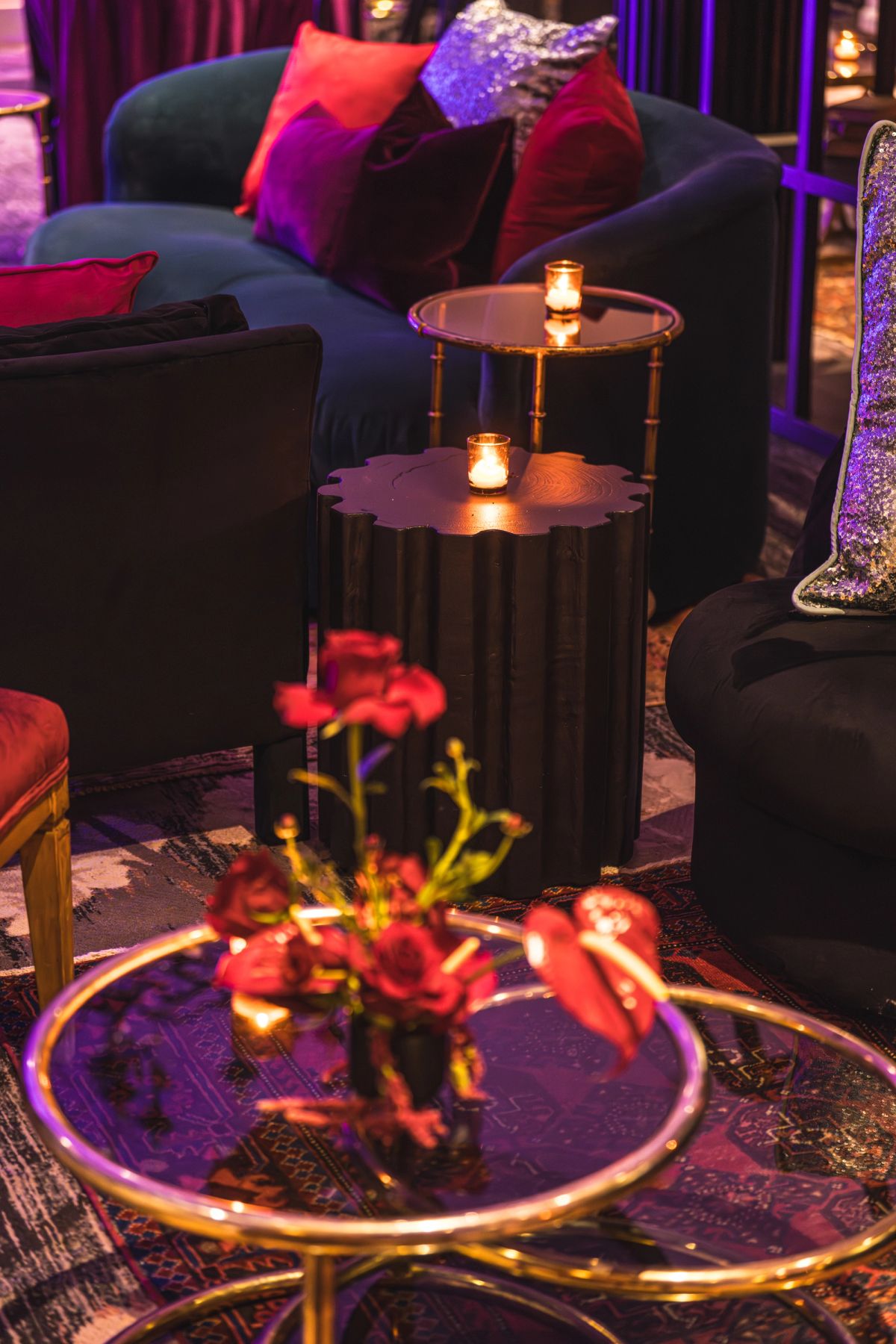 Elegant lounge seating with dark sofas, colorful pillows, glass coffee table with red roses, and warm ambient.