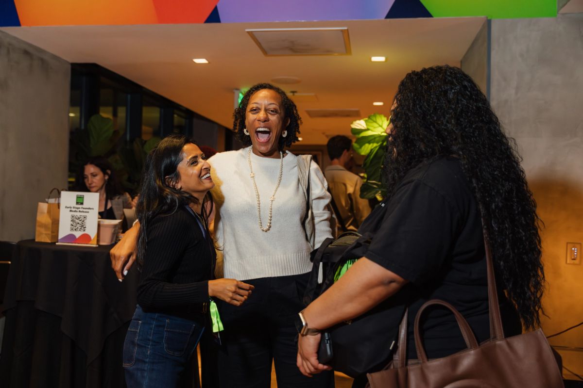 Three women embrace and laugh together in a modern conference venue lobby with colorful ceiling design.