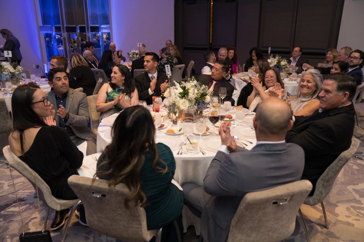 Formal dinner event with guests seated at round tables with white linens and floral centerpieces, applauding.