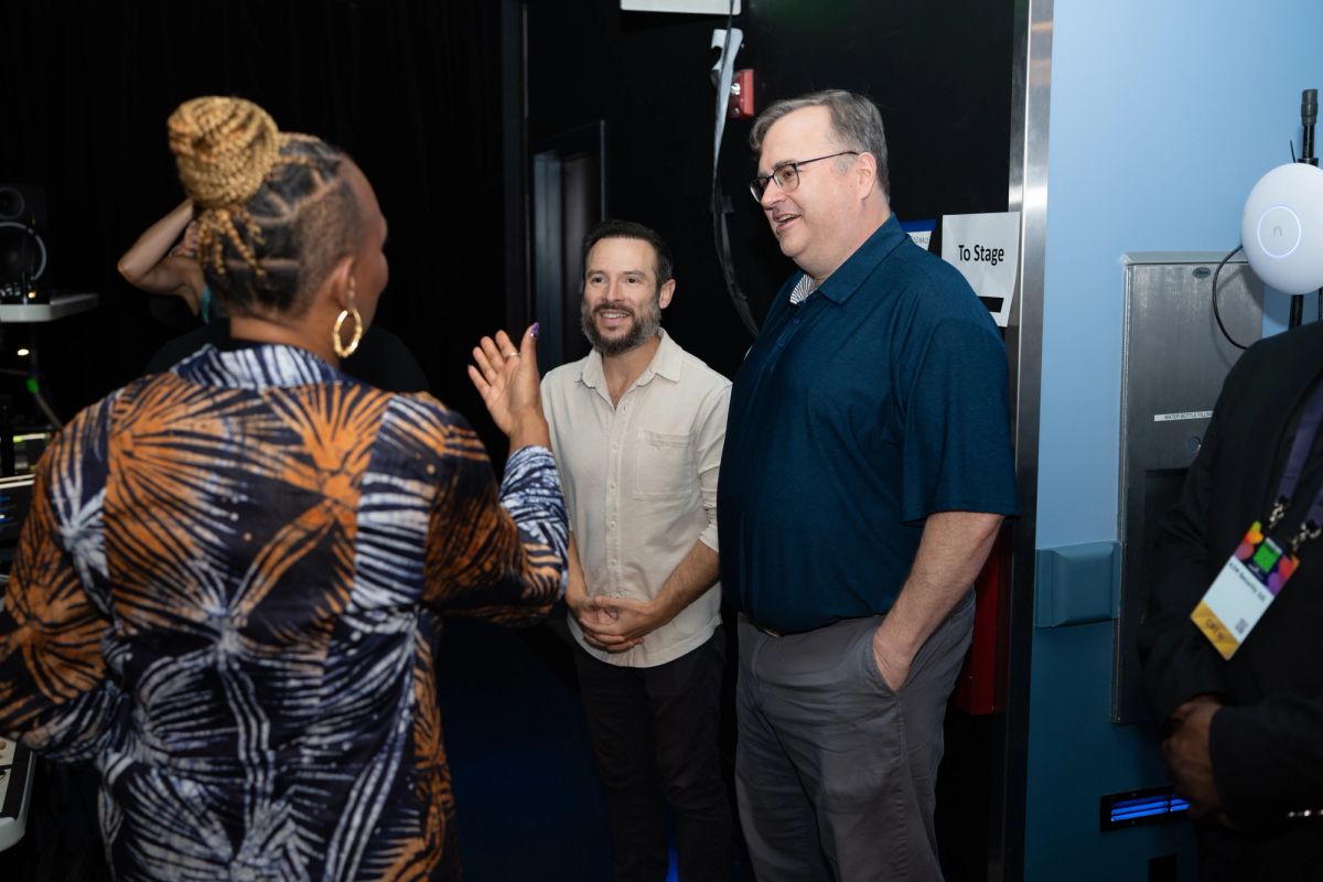 Three people networking backstage at a conference venue, with one person gesturing while speaking to two men.